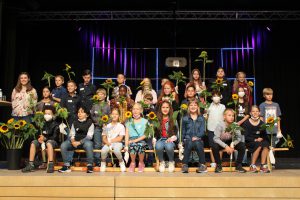 Eine Gruppe von Kindern und Jugendlichen steht und sitzt auf einer Bühne, umgeben von Sonnenblumen. Sie sind in verschiedenen Kleidungsstilen gekleidet und zeigen eine fröhliche Stimmung. Im Hintergrund sind Lichter und Dekorationen sichtbar, die eine festliche Atmosphäre schaffen.