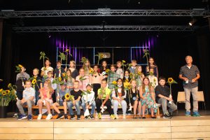 Gruppenfoto von Schülern auf einer Bühne. Die Kinder halten Sonnenblumen in den Händen und sind in verschiedenen Kleidungsstilen gekleidet. Im Hintergrund sind Bühnenlichter sichtbar. Ein Erwachsener steht rechts neben den Kindern.