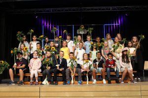Gruppe von Kindern und Jugendlichen, die auf einer Bühne sitzen und Sonnenblumen in den Händen halten. Sie stehen in mehreren Reihen, einige tragen einheitliche T-Shirts. Der Hintergrund ist dunkel mit bunten Lichtern.
