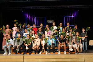 Eine Gruppe von Schülern steht auf einer Bühne, lächelt und hält Sonnenblumen in den Händen. Sie sind in mehreren Reihen angeordnet und tragen verschiedene Kleidungsstile. Im Hintergrund sind lila Beleuchtungen und ein Bühnenaufbau zu sehen.
