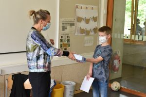 Eine Frau und ein Junge stehen sich gegenüber, beide tragen Mund-Nasen-Schutz. Die Frau reicht dem Jungen ein kleines Paket, während er ein Blatt Papier in der Hand hält. Im Hintergrund sind einige Plakate und ein Fenster sichtbar. Der Raum wirkt hell und freundlich.