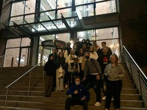 Eine Gruppe von etwa 20 Personen steht auf den Treppen vor einem modernen Gebäude mit großen Fenstern. Die Menschen tragen verschiedene Kleidung und einige haben Masken auf. Die Szene ist abends, mit Licht von innen, das die Umgebung erhellt.