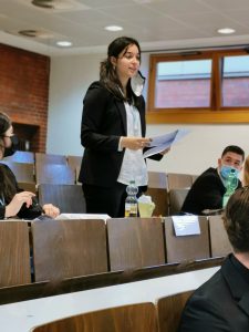 Eine Frau steht in einem Seminarraum und spricht. Sie trägt einen schwarzen Anzug und hält Unterlagen in der Hand. Im Hintergrund sind mehrere Personen in Sitzreihen zu sehen, einige tragen Masken. Der Raum hat Fenster, durch die Tageslicht einfällt.