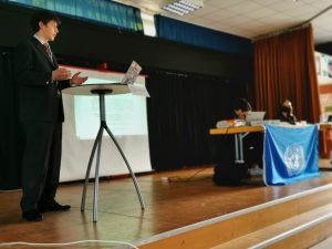 Ein junger Mann in Anzug steht an einem Tisch und präsentiert vor einem Publikum. Im Hintergrund ist eine Projektionsleinwand mit Text zu sehen. An einem Tisch sitzt eine Person, die auf einen Laptop schaut, während eine weitere Person neben einer Fahne der Vereinten Nationen steht. Der Raum ist gut beleuchtet und hat eine Bühne.