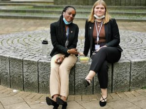 Zwei Frauen sitzen auf einem runden Steinpflaster. Die Frau links trägt eine schwarze Jacke und helle Hosen, die Frau rechts trägt ebenfalls eine schwarze Jacke und schwarze Hosen. Beide haben einen Ausweis um den Hals und lächeln in die Kamera. Im Hintergrund ist eine grüne, leicht strukturierte Fläche sichtbar.
