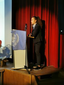 Eine Person in formeller Kleidung steht an einem Rednerpult mit einer Flagge der Vereinten Nationen. Der Hintergrund ist rot und zeigt eine Präsentation. Die Person spricht in ein Mikrofon und hat lange, lockige Haare.