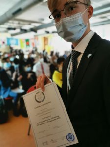Ein junger Mann in einem Anzug und mit einer Maske hält ein Zertifikat in der Hand. Im Hintergrund sind viele Menschen zu sehen, die an einer Veranstaltung teilnehmen. Die Szene vermittelt eine festliche Atmosphäre.