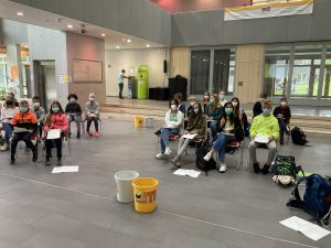 Eine Gruppe von Personen sitzt in einem großen Raum, während sie Abstand halten. Alle tragen Masken. Einige Teilnehmer haben Notizblätter und Stifte bei sich. Im Vordergrund stehen zwei Eimer. Im Hintergrund sind große Fenster und eine Bühne sichtbar.