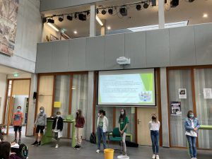 Eine Gruppe von Kindern steht in einem großen Raum, umgeben von Fenstern und einer Präsentationsleinwand. Die Kinder tragen Masken und stehen in einer Reihe. Auf der Leinwand ist ein Text oder eine Präsentation sichtbar. Der Raum hat eine moderne Architektur mit Holz- und Glasdetails.