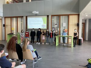 Eine Gruppe von zehn Personen steht auf einer Bühne in einem modernen Raum. Sie tragen Masken und stehen nebeneinander, während das Publikum, bestehend aus mehreren Personen, in Stühlen davor sitzt. Im Hintergrund ist ein Präsentationsbildschirm zu sehen, auf dem Informationen angezeigt werden. Der Raum hat große Fenster und eine helle, freundliche Atmosphäre.