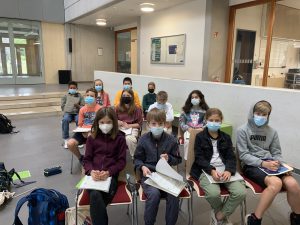 Eine Gruppe von Kindern sitzt in einem hellen Raum auf Stühlen. Alle tragen Mund-Nasen-Schutzmasken. Die Kinder halten Bücher oder Notizblätter und blicken nach vorne. Im Hintergrund sind große Fenster und einige Türen zu sehen.