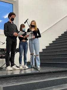 Drei Personen stehen auf einer Bühne, während sie ein Mikrofon benutzen. Zwei der Personen tragen schwarze Kleidung und eine Person hat eine Jeans und ein schwarzes Oberteil an. Alle tragen Masken. Im Hintergrund sind Treppen zu sehen, und es scheint Tageslicht durch ein Fenster zu kommen.