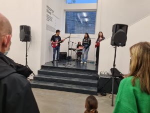 Eine Band mit vier Jugendlichen spielt auf einer kleinen Bühne in einem Ausstellungsraum. Zwei der Musiker spielen E-Gitarre, einer sitzt am Keyboard und eine weitere Musikerin steht neben ihnen. Im Vordergrund sind einige Zuschauer zu sehen, die der Aufführung aufmerksam folgen. Die Wände sind hell und der Raum wirkt modern und offen.