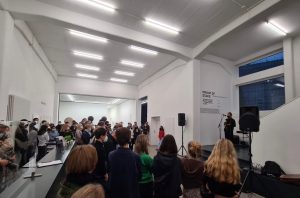 Eine Gruppe von Menschen steht in einem hellen, modernen Raum mit weißen Wänden und großen Fenstern. Im Vordergrund ist ein Redner am Mikrofon zu sehen, während das Publikum aufmerksam zuhört. Die Anwesenden sind unterschiedlich gekleidet und stehen in einer lockeren Ansammlung. Im Hintergrund sind einige Gegenstände und Lautsprecher sichtbar.
