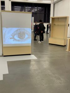 Ein Ausstellungsraum mit einem Projektor, der ein großes Augenbild an eine Wand strahlt. Im Vordergrund stehen zwei Ausstellungsvitrinen, während Besucher im Hintergrund stehen und die Ausstellung betrachten. Der Boden ist grau und die Wände sind hell.