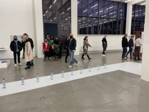 Eine Gruppe von Menschen besucht eine Ausstellung in einem hellen Raum mit großen Fenstern. Im Vordergrund ist eine Reihe von Wasserflaschen auf einem weißen Streifen zu sehen. Die Besucher sind in verschiedenen Kleidungsstücken gekleidet und bewegen sich durch den Raum, während einige miteinander sprechen und andere die Ausstellung betrachten.