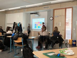 Ein Klassenzimmer mit Schülern, die auf einen Vortrag achten. Im Vordergrund sitzen mehrere Schüler an Tischen, während vorne an der Wand ein Projektor eine Präsentation zeigt. Eine Person steht vor der Präsentation und spricht. An der Wand hängt eine Uhr, und verschiedene Gegenstände sind auf den Tischen verteilt.
