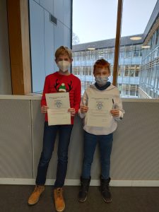 Zwei Kinder stehen nebeneinander und halten jeweils ein Zertifikat in den Händen. Beide tragen Mund-Nasen-Schutzmasken. Das Kind links trägt einen roten Pullover mit einem Rentiermotiv, das Kind rechts trägt einen weißen Pullover. Im Hintergrund ist ein moderner Gebäude mit großen Fenstern zu sehen.