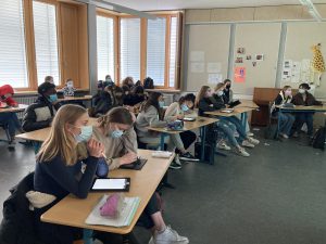 Eine Klasse mit Schülern, die in einem Raum mit großen Fenstern sitzen. Die meisten Schüler tragen Mund-Nasen-Schutz und nutzen Tablets oder Notizblätter. An der Wand sind einige Bilder und eine Zeichnung einer Giraffe zu sehen.