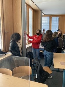 Eine Gruppe von Schülern in einem Klassenraum. Einige stehen um einen Tisch herum, während eine Schülerin in roter Jacke an einer Wand steht und etwas erklärt. Alle tragen Masken. Der Raum hat große Fenster und eine helle, freundliche Atmosphäre.
