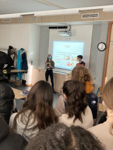 Zwei Personen stehen vor einer Gruppe von Menschen in einem Klassenraum. Eine Person hält ein Blatt Papier oder ein Buch in der Hand und spricht, während die andere Person neben ihr steht. Im Hintergrund ist ein Bildschirm mit einer Präsentation sichtbar. Die Anwesenden schauen in Richtung der Sprecher. An der Wand hängt eine Uhr und es sind einige Jacken an einem Kleiderständer zu sehen.