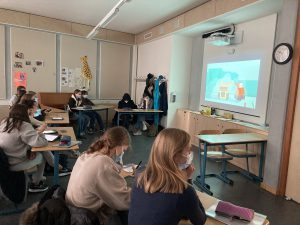 Eine Gruppe von Schülern sitzt in einem Klassenzimmer und schaut auf einen Bildschirm, auf dem eine Animation zu sehen ist. Die Schüler tragen Masken und sind mit Notizmaterialien beschäftigt. Im Hintergrund sind weitere Schüler und Unterrichtsmaterialien sichtbar. An der Wand hängt eine Uhr und es gibt eine Giraffenfigur, die zur Dekoration dient.