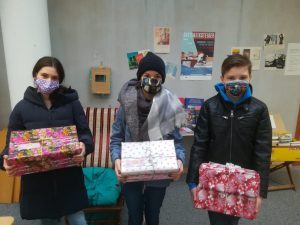 Drei Kinder stehen in einem Raum und halten jeweils ein Geschenk in den Händen. Sie tragen Masken und warme Kleidung. Im Hintergrund sind ein Stuhl und verschiedene Plakate an der Wand zu sehen.