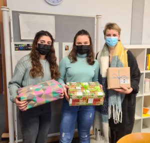 Drei Personen stehen in einem Raum und halten Geschenke in den Händen. Alle tragen Mund-Nasen-Schutz. Zwei Frauen stehen links und in der Mitte, während die dritte Frau rechts steht und ein Geschenk in einer neutralen Verpackung hält. Der Hintergrund zeigt eine Pinnwand und Regale.