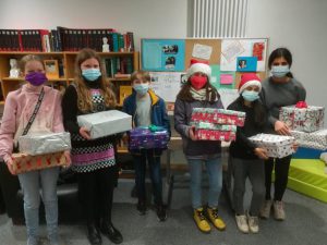 Sechs Kinder stehen in einem Raum, alle tragen Masken. Sie halten Geschenke in den Händen, einige sind in bunte Verpackungen gehüllt, andere in silbernes oder rotes Papier. Im Hintergrund sind Regale mit Büchern und ein Pinnwand mit verschiedenen Informationen zu sehen. Einige Kinder tragen Weihnachtsmützen.
