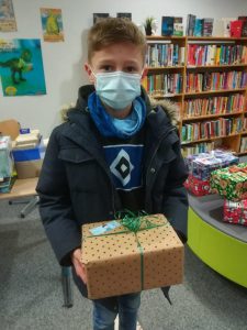 Ein Junge mit einem Mund-Nasen-Schutz hält ein Geschenk in seinen Händen. Er trägt eine dunkle Jacke und einen blauen Schal. Im Hintergrund sind Regale mit Büchern und weitere Geschenke zu sehen.