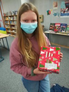 Ein Mädchen mit langen, blonden Haaren trägt eine blaue Maske und hält ein bunt verpacktes Geschenk in den Händen. Der Hintergrund zeigt eine Bibliothek mit Regalen voll Bücher. Das Geschenk ist in rotem Papier mit Weihnachtsbaum-Motiven verpackt.