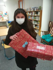 Ein Mädchen mit langen Haaren trägt eine Maske und hält zwei schön verpackte Geschenke in rotem Geschenkpapier. Im Hintergrund sind Bücherregale und ein Stuhl zu sehen, was auf eine gemütliche Umgebung hindeutet.