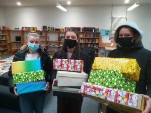Drei Personen stehen in einem Raum mit Bücherregalen im Hintergrund. Sie tragen Gesichtsmasken und halten mehrere bunt verpackte Geschenke in den Händen. Die Geschenke sind in verschiedenen Farben und mit unterschiedlichen Mustern versehen.