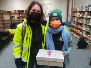 Zwei Personen stehen in einem Raum mit Bücherregalen im Hintergrund. Die linke Person trägt eine leuchtend gelbe Jacke und einen schwarzen Mund-Nasen-Schutz. Die rechte Person trägt eine grüne Mütze und einen orangefarbenen Mund-Nasen-Schutz. Beide lächeln und halten gemeinsam eine Geschenkschachtel mit rotem Band. Der Boden ist mit Teppich ausgelegt und es sind einige Möbel im Hintergrund sichtbar.