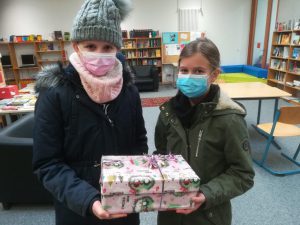 Zwei Mädchen stehen in einer Bibliothek und halten ein Geschenk in den Händen. Beide tragen Mund-Nasen-Schutzmasken. Das Geschenk ist bunt verpackt und hat eine Schleife. Im Hintergrund sind Regale mit Büchern zu sehen.
