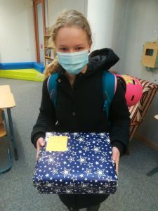 Ein Mädchen trägt einen Rucksack und hält ein blaues, mit Sternen bedrucktes Geschenk in den Händen. Sie trägt eine Maske und steht in einem Raum mit Möbeln im Hintergrund.