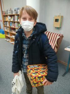 Ein junger Junge mit blonden Haaren trägt eine Maske und steht in einem Raum mit Bücherregalen. Er hat einen schwarzen Wintermantel und ein kariertes Hemd an. In seiner rechten Hand hält er ein bunt verpacktes Geschenk, während er in der linken Hand eine weiße Tragetasche hält.