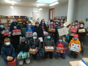 Eine Gruppe von Kindern und Jugendlichen steht in einem Raum, in dem Bücherregale sichtbar sind. Alle tragen Masken und halten liebevoll verpackte Geschenke in den Händen. Die Geschenke sind unterschiedlich groß und in buntem Geschenkpapier eingewickelt. Der Raum hat helle Wände und viel Tageslicht. Es gibt eine freundliche, gemeinschaftliche Atmosphäre.