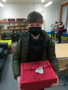 Ein junger Mann mit einem schwarzen Mund-Nasen-Schutz hält ein rotes Geschenkpaket in seinen Händen. Im Hintergrund sind Regale mit Büchern und weitere Personen zu sehen, die miteinander interagieren. Der Raum ist hell erleuchtet und hat eine freundliche, einladende Atmosphäre.