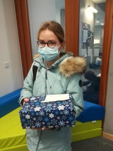 Ein Mädchen mit einer Brille und einem Mund-Nasen-Schutz trägt eine blaue Winterjacke und hält ein hübsch verpacktes Geschenk in den Händen. Das Geschenk ist mit blauen und weißen Schneeflockenmotiven dekoriert. Im Hintergrund sind Teile eines Raumes mit Möbeln und einem farbigen Sitzbereich zu sehen.