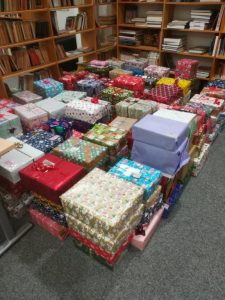 Eine Vielzahl von bunt verpackten Geschenken in unterschiedlichen Größen, die auf dem Boden gestapelt sind. Im Hintergrund sind Regale mit Ordnern und Bücher zu sehen. Das Bild vermittelt eine festliche Atmosphäre.