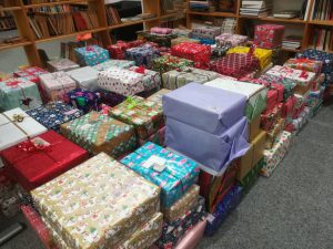 Eine große Ansammlung von bunt verpackten Geschenken in verschiedenen Farben und Mustern, die auf dem Boden liegen. Im Hintergrund sind Regale mit Büchern sichtbar.