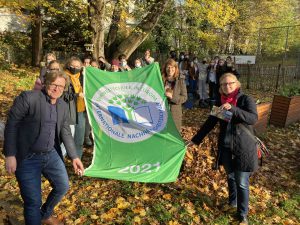 Eine Gruppe von Menschen steht im Freien in einem herbstlichen Park. Im Vordergrund halten zwei Personen eine große grüne Flagge mit einem Logo und der Jahreszahl 2021. Im Hintergrund sind weitere Personen zu sehen, die ebenfalls an der Veranstaltung teilnehmen. Die Umgebung ist von buntem Laub geprägt.