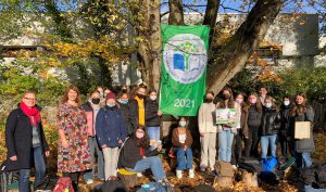 Gruppe von Menschen, die unter einem Baum stehen und eine grüne Fahne mit dem Jahr 2021 halten. Die Teilnehmer tragen Masken und stehen in einem parkähnlichen Umfeld mit herbstlichem Laub. Einige halten Plakate oder andere Gegenstände.