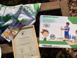 Auf dem Bild liegen ein grünes und weißes Tuch, ein Bausatz für eine Windkraftanlage, eine Urkunde sowie ein Informationsflyer auf einem Holzuntergrund. Die Urkunde ist in einem Rahmen, und der Bausatz zeigt ein Kind, das damit spielt. Die Umgebung ist mit herbstlichen Blättern bedeckt.