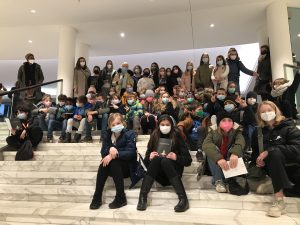 Eine große Gruppe von Menschen sitzt und steht auf einer Treppe in einem hellen, modernen Gebäude. Alle tragen Masken. Die Gruppe besteht aus Kindern und Erwachsenen, die in verschiedenen Positionen und Posen angeordnet sind. Einige Personen sitzen auf den Stufen, während andere im Hintergrund stehen.
