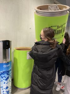 Eine Person in einem schwarzen Mantel steht neben zwei großen, grünen Behältern. Die Person greift nach dem oberen Behälter, der mit der Aufschrift „Dein Stand Up“ dekoriert ist. Im Hintergrund ist ein Wasserspender sichtbar. Die Umgebung wirkt modern und hell.