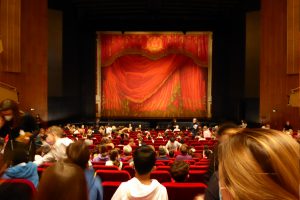 Blick auf ein Theaterpublikum, das auf eine große rote Bühnenvorhang wartet. Die Zuschauer sitzen in roten Sitzreihen, während der Vorhang mit kunstvollen Mustern im Hintergrund sichtbar ist.