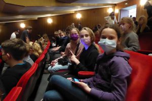 Drei Jugendliche sitzen in einem Theater mit rotem Plüschsitz. Sie tragen Masken und schauen in die Kamera. Im Hintergrund sind weitere Personen zu sehen, die ebenfalls im Zuschauerraum sitzen. Die Beleuchtung ist warm und gemütlich.