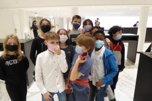 Gruppe von zehn Kindern und Jugendlichen in einem modernen Innenraum, alle tragen Gesichtsmasken. Sie stehen eng beieinander und lächeln, einige posieren spielerisch. Der Hintergrund zeigt eine helle, minimalistische Umgebung mit einem Empfangstresen.